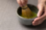Hands whisking green matcha in a rustic ceramic bowl on a woven gray mat. Relaxed, focused mood. No visible text.