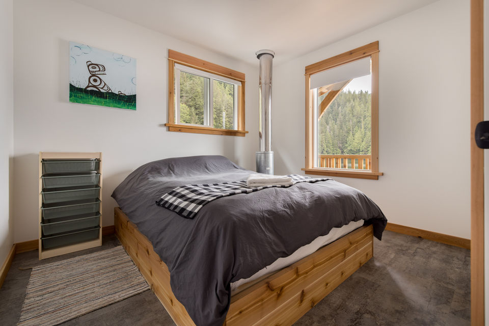 Cozy private bedroom inside the Khutzeymateen Wilderness Lodge with a queen-size bed, large windows with forest views, and rustic decor.