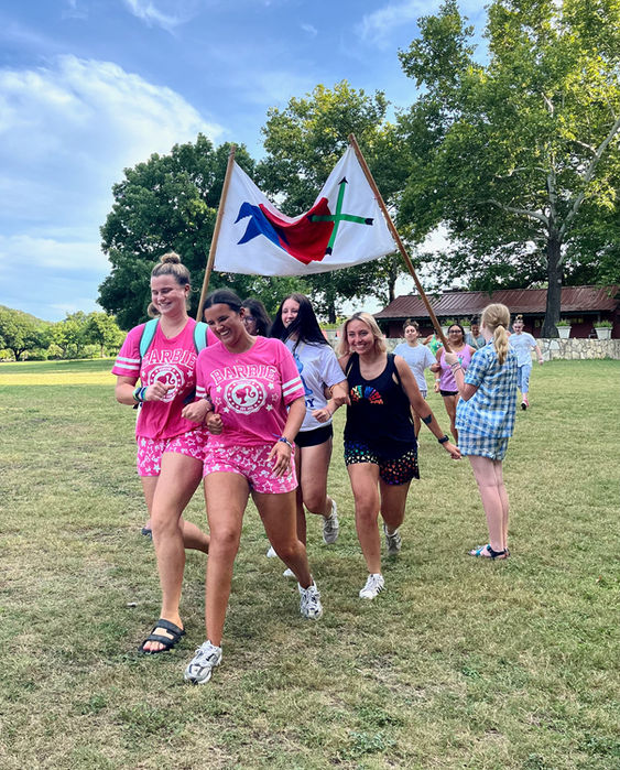 Group of girls in a field jogging under a team flag that is held up by counselors.