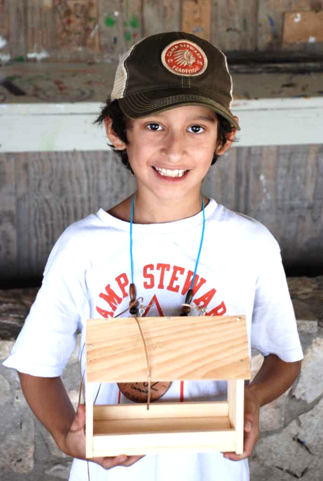 Boy wearing a Camp Stewart t-shirt and a baseball cap holding a wooden bird feeder that he made