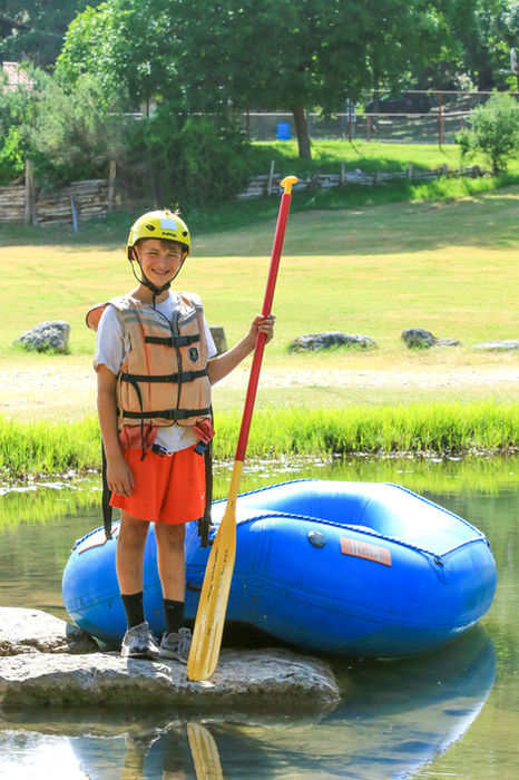 Boy standing in front of an inflatable kayak in the river, holding an oar