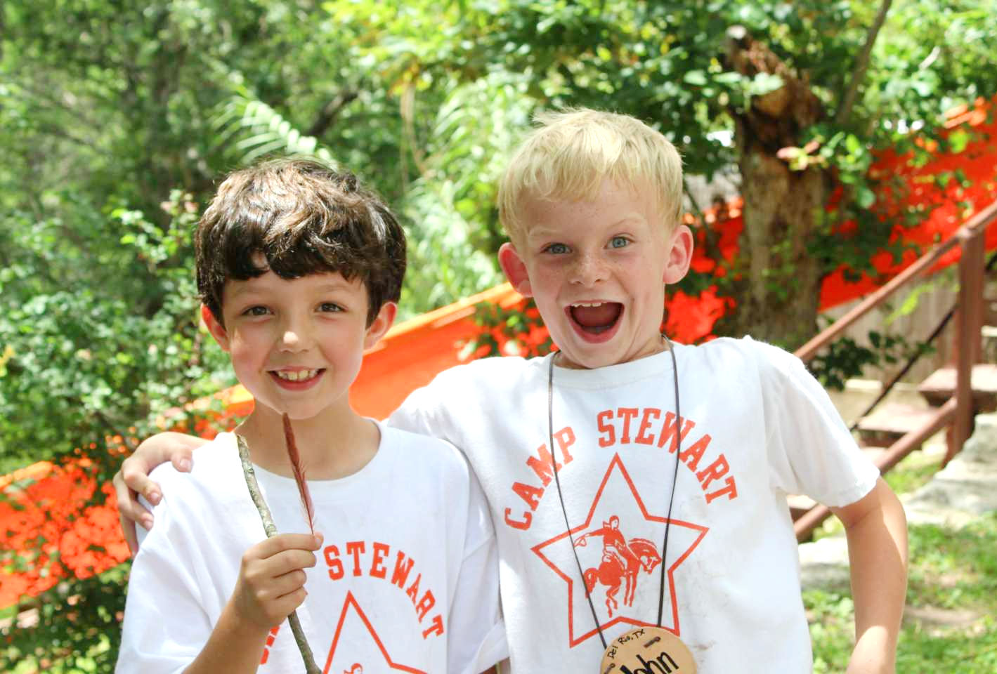 Two young boys, standing outdoors, smiling, wearing Camp Stewart t-shirts