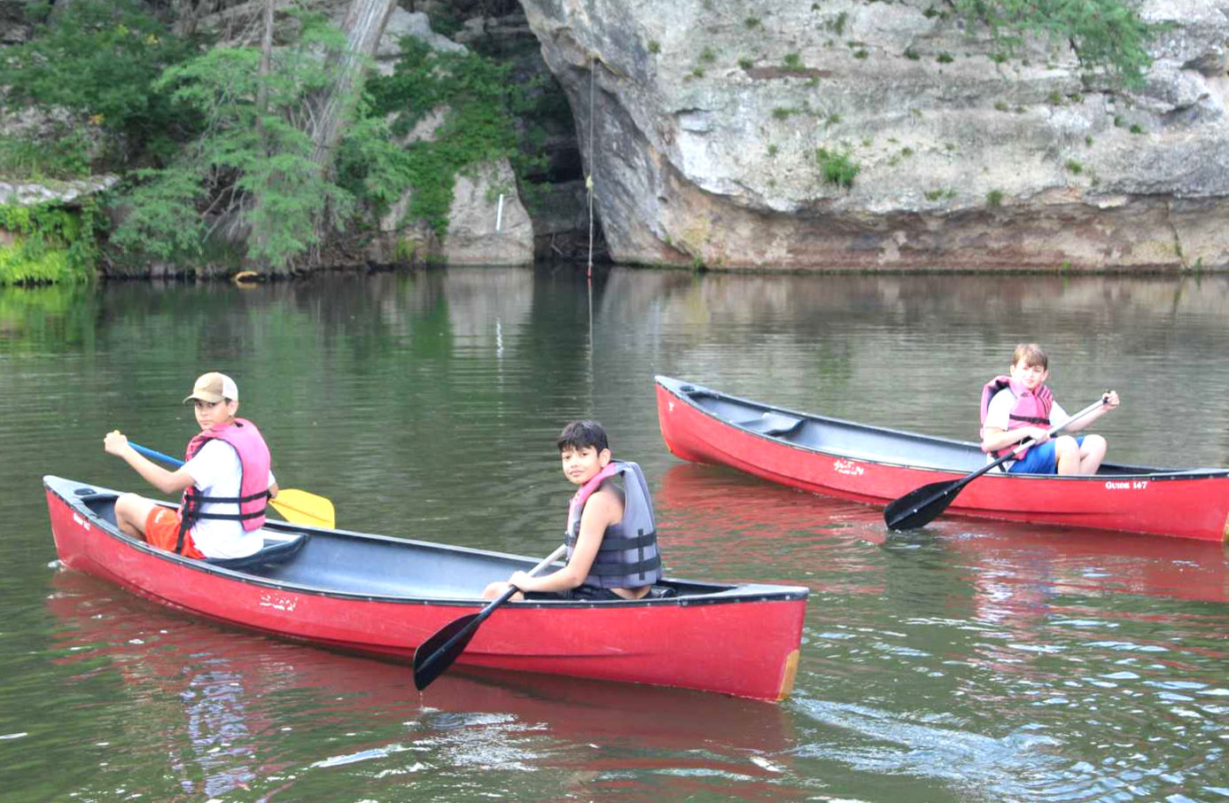 Two boys in one red canoe, one boy in another, paddling on a river