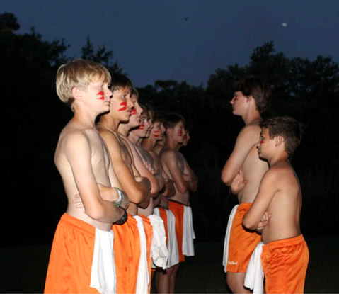 Two rows of boys, facing each other, all shirtless wearing orange shorts and white towels, outdoors at night