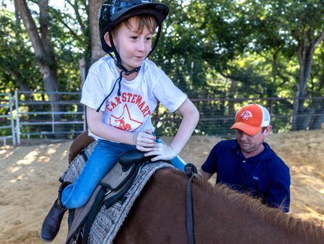 Young boy wearing a Camp Stewart t-shirt and equestrian helmet sitting on a brown horse