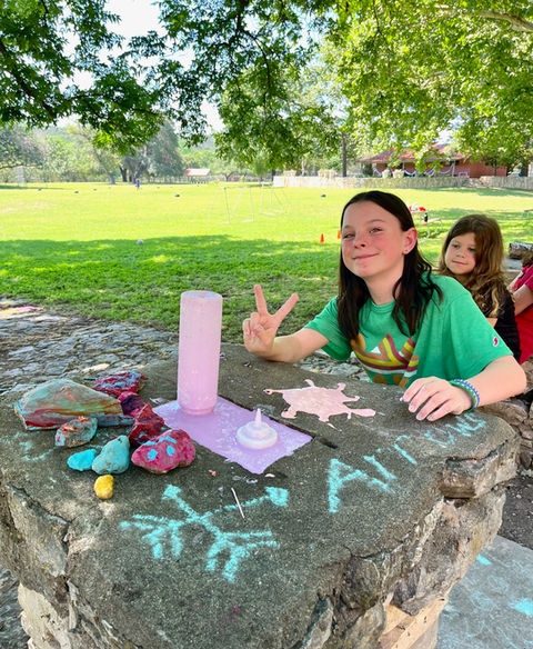 Young girl outdoors, standing at a rock fence, making the peace sign and smiling at the camera. There are candles and painted rocks in front of her on the rock fence.