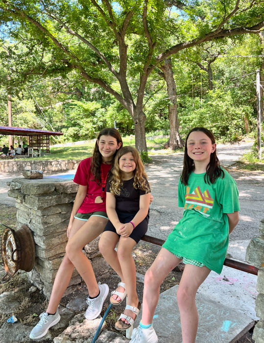 Three girls at Heart O' the HIlls girls camp outdoors, sitting on a rail, looking at the camera and smiling.