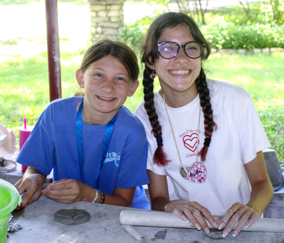 Two girls sitting outdoors at an arts and crafts table, looking at the camera and smiling.