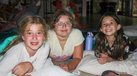 Three young girls at a camp slumber party, laying on the ground propped up on their elbows, looking at the camera, smiling.