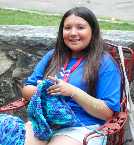 Girl in a blue t-shirt who is knitting with bright, multi-colored yarn.