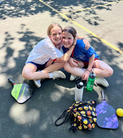 Two girls at Heart O' the Hills girls camp sitting on the ground hugging each other, looking at the camera and giggling, with pickleball paddles and a ball on the ground.