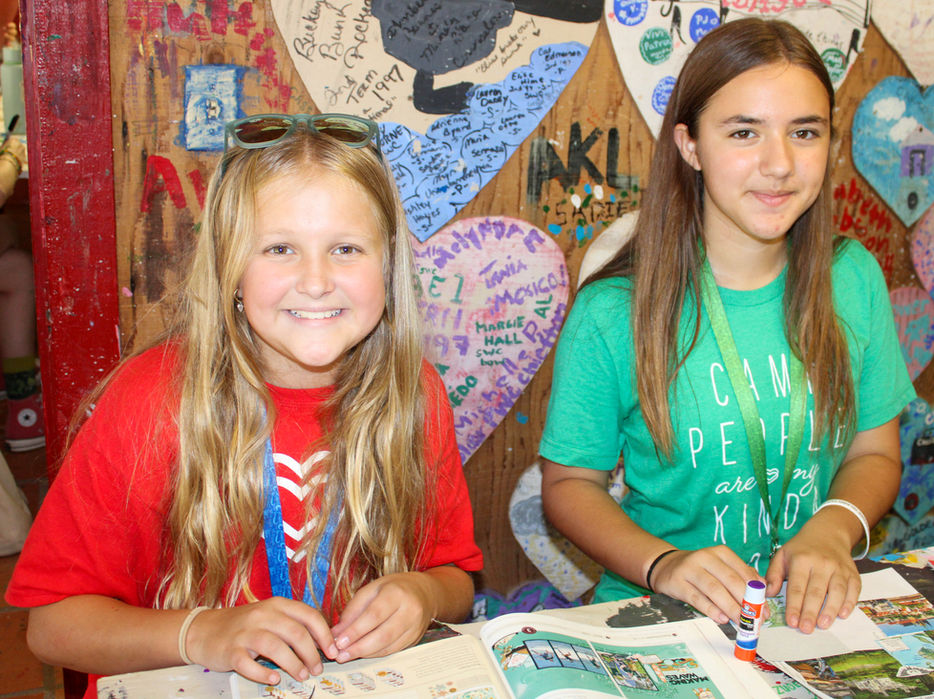Two girls sitting at an arts and crafts table, looking at the camera and smiling.