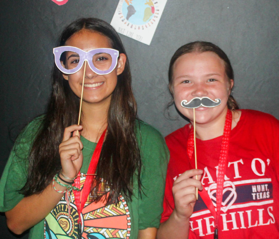 Two girls looking at the camera and smiling. One is holding a 'mask' of large eye glass frames in front of her face. The other is holding a 'mask' of a mustache in front of the face.