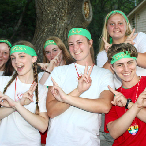 Six older girls wearing t-xhirts and green headbands, looking at the camera and smiling, and making peace sign like hand gestures.