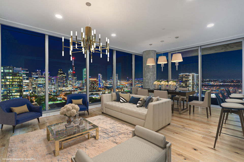 DallasPro luxury high-rise real estate photography taken at night. A bright, open-concept living and dining area features floor-to-ceiling windows that provide a crystal-clear, reflection-free panoramic view of the illuminated downtown Dallas skyline. The interior is flawlessly exposed, highlighting a modern gold chandelier, light wood floors, modern seating, and sophisticated virtual staging, perfectly contrasting with the dark night sky and bright city lights.