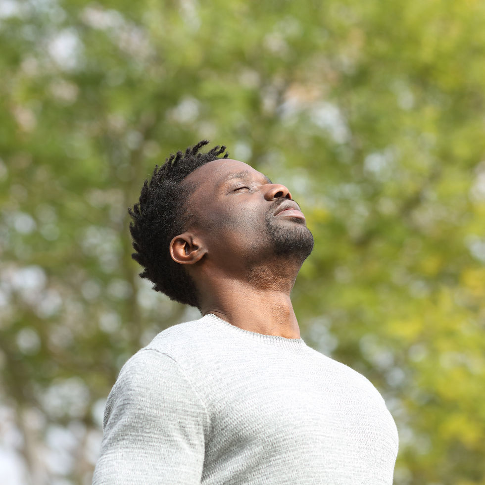 Man in a light sweater stands outdoors with eyes closed, appearing relaxed. Green trees blur in the background, conveying a peaceful mood.