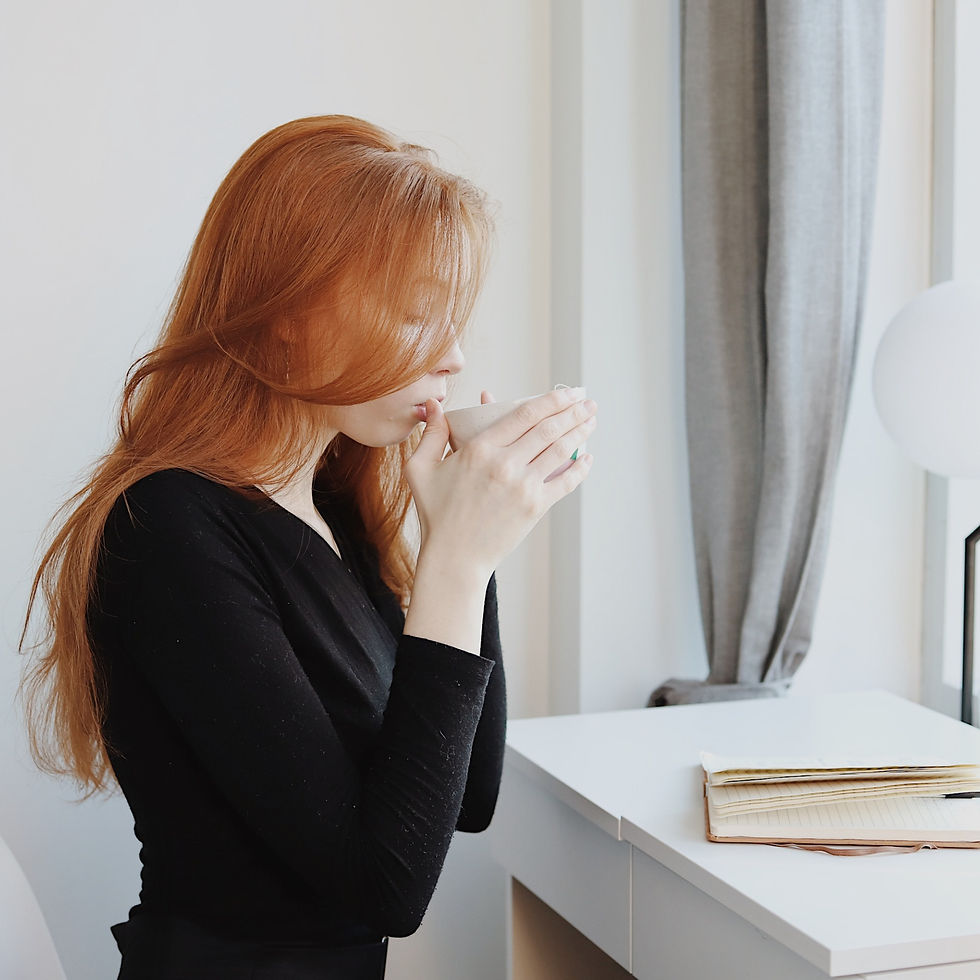 Red-haired woman in black sweater sips coffee at a white desk by a window. Notebooks sit on the desk. Bright, relaxed atmosphere.