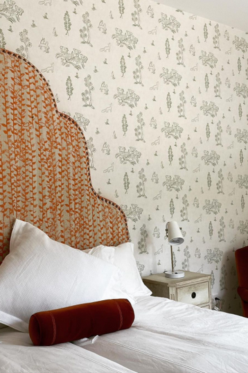 white and light green flora and  fauna patterned wallpaper next to a high scalloped headboard with white linens and scarlet accents.
