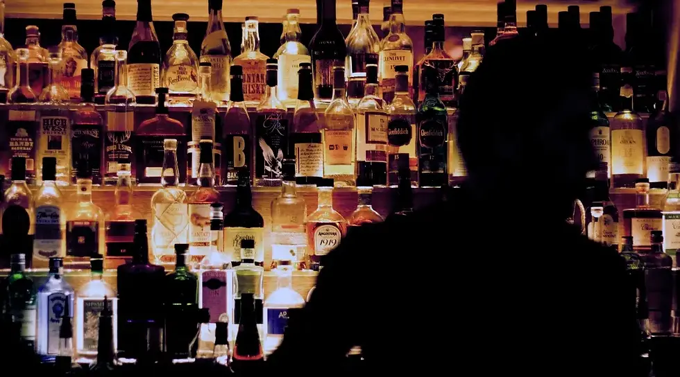 Silhueta de um homem em frente a um bar, ao fundo garrafas de bebidas alcoólicas em prateleiras, insinuando a crise de intoxicação por metanol