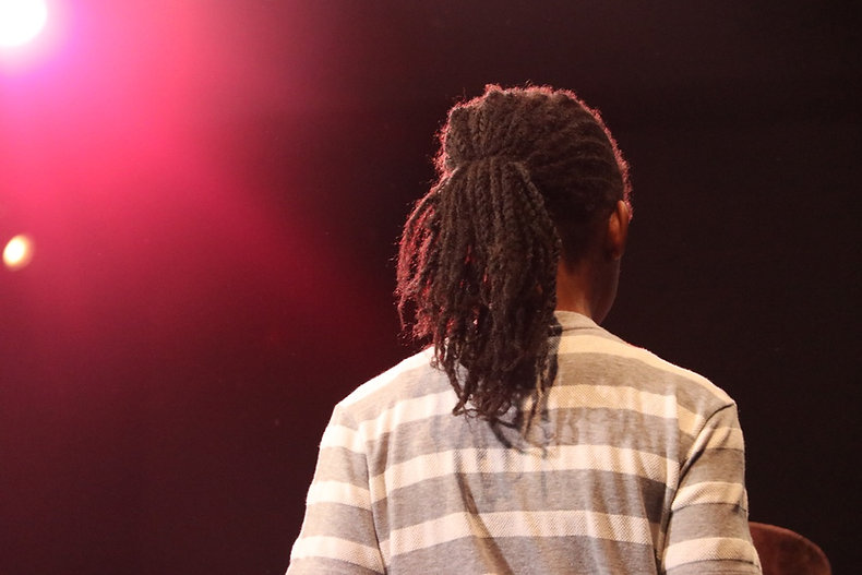 The back of a young Black person with locks in a ponytail and a grey and white striped shirt on stage with warm red and yellow lights.