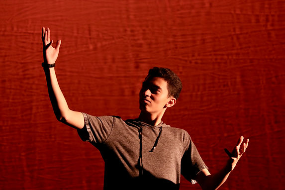 A student stands onstage in front of a red background holding up one hand and looking at that hand. The student has tan skin, dark short hair, and wears a grey short sleeved hoodie.