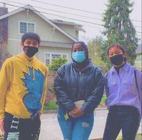 Three young people of color in face masks stand outside looking at the camera in front of a house and trees.