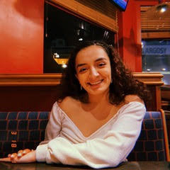 Sara, long curly black hair wearing a long sleeve white shirt, sits in the booth of restaurant smiling