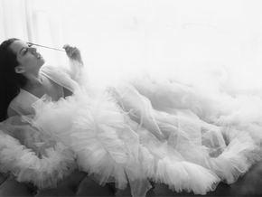 Black and white bridal boudoir portrait of woman in flowing tulle gown reclining on sofa near window light.