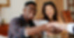 A male and a female sitting on one side of the table. The male is extending his hand across the table to shake the hand of another individual who is only partially in the frame.
