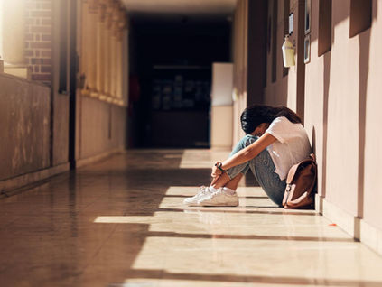 Young woman sits alone in a sunlit hallway, knees hugged to chest, backpack beside her, looking sad and withdrawn.