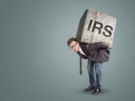 Man in suit and glasses strains under a giant stone block labeled IRS against a plain gray background.