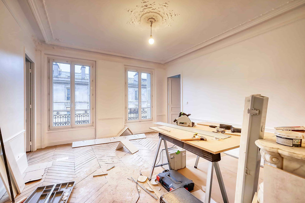 Renovation work in a bright empty room with parquet flooring, tools on a workbench, two windows, and a single hanging bulb.
