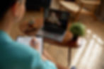Person taking notes during a video call on a laptop in a cozy living room, with a glass, glasses, and potted plant on the table
