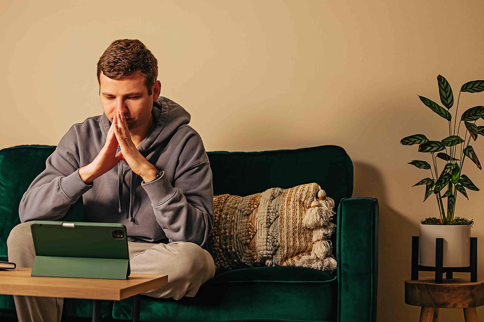 Man in gray hoodie sits on a green couch, hands clasped in prayer, looking at a tablet in a cozy room with a potted plant.