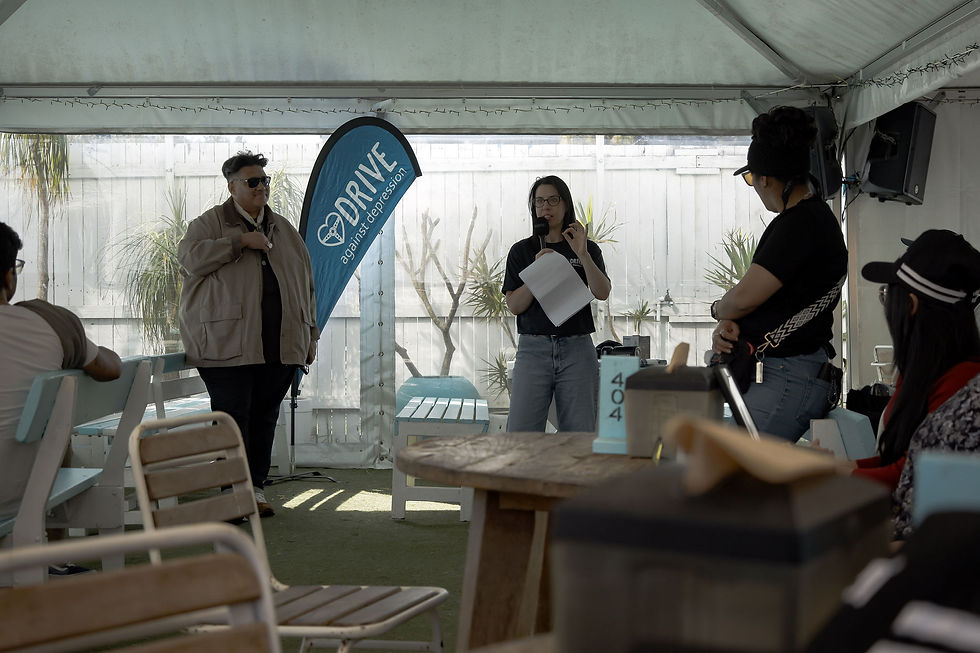 People listen to a speaker under a tent beside a blue DRIVE against depression banner during an outdoor community talk.
