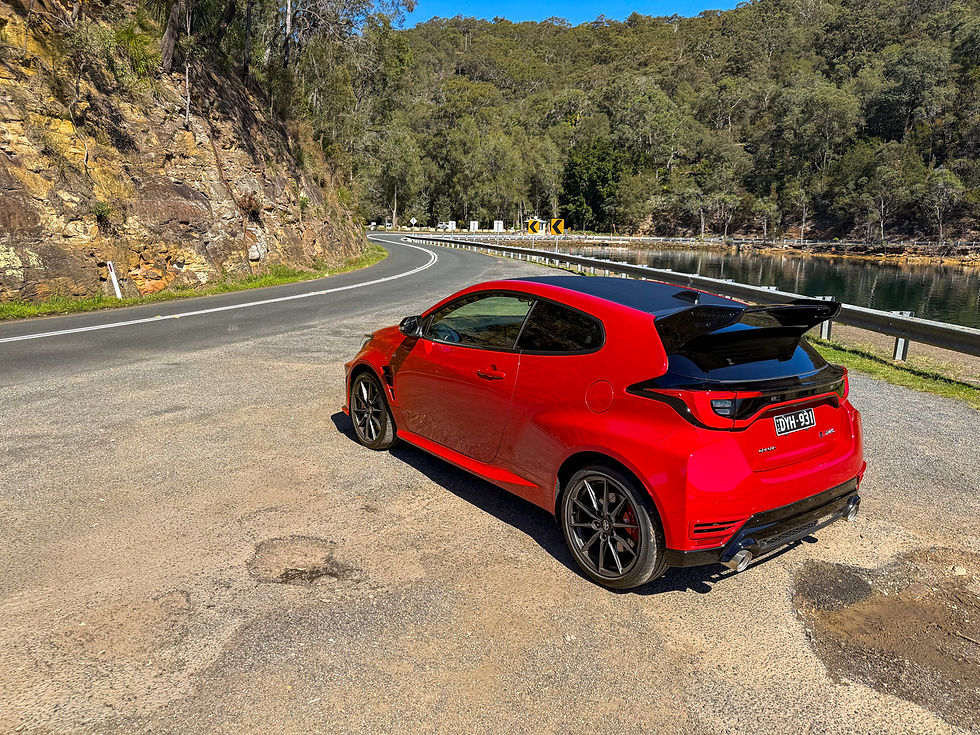 Red hatchback parked on a winding mountain road beside water, with forested cliffs and clear blue sky on a sunny day.