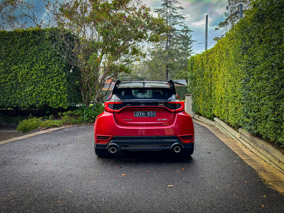 Red Toyota GR Yaris parked on a leafy driveway between tall hedges, rear view, with plate DYH-931.