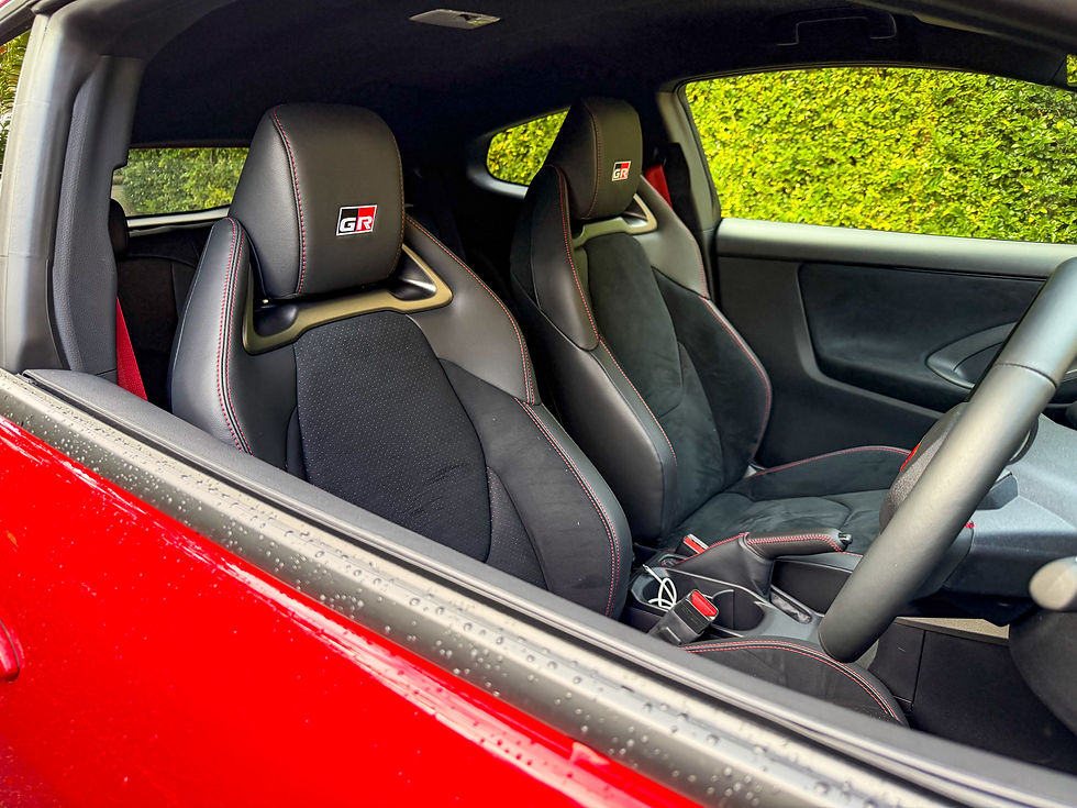 Red sports car interior with black GR bucket seats and manual shifter, parked beside a bright green hedge.