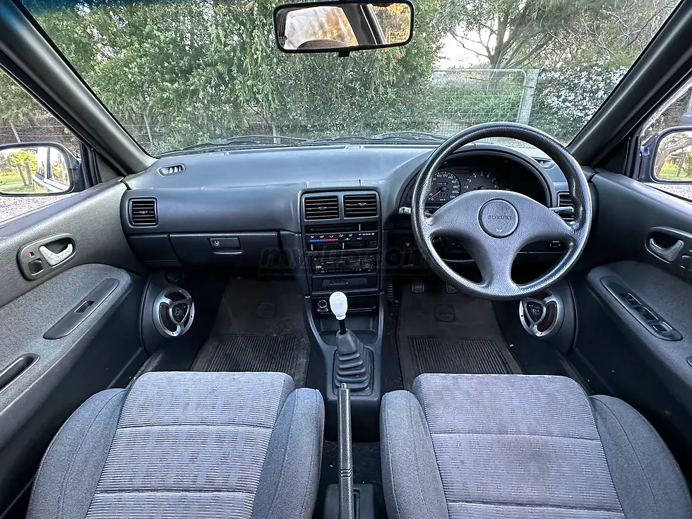 Right-hand-drive Suzuki interior with manual gear shift, gray seats, and trees visible through the windshield.