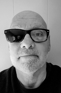 A black and white photo of Author Wilder Sanderline with stubble, glasses in front of a grey wall