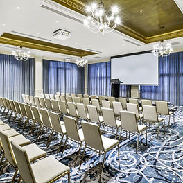A modern, well-lit conference room at The Playford Hotel in Adelaide featuring theater-style seating, elegant chandeliers, blue-patterned carpeting, and a large projection screen, perfect for professional events.