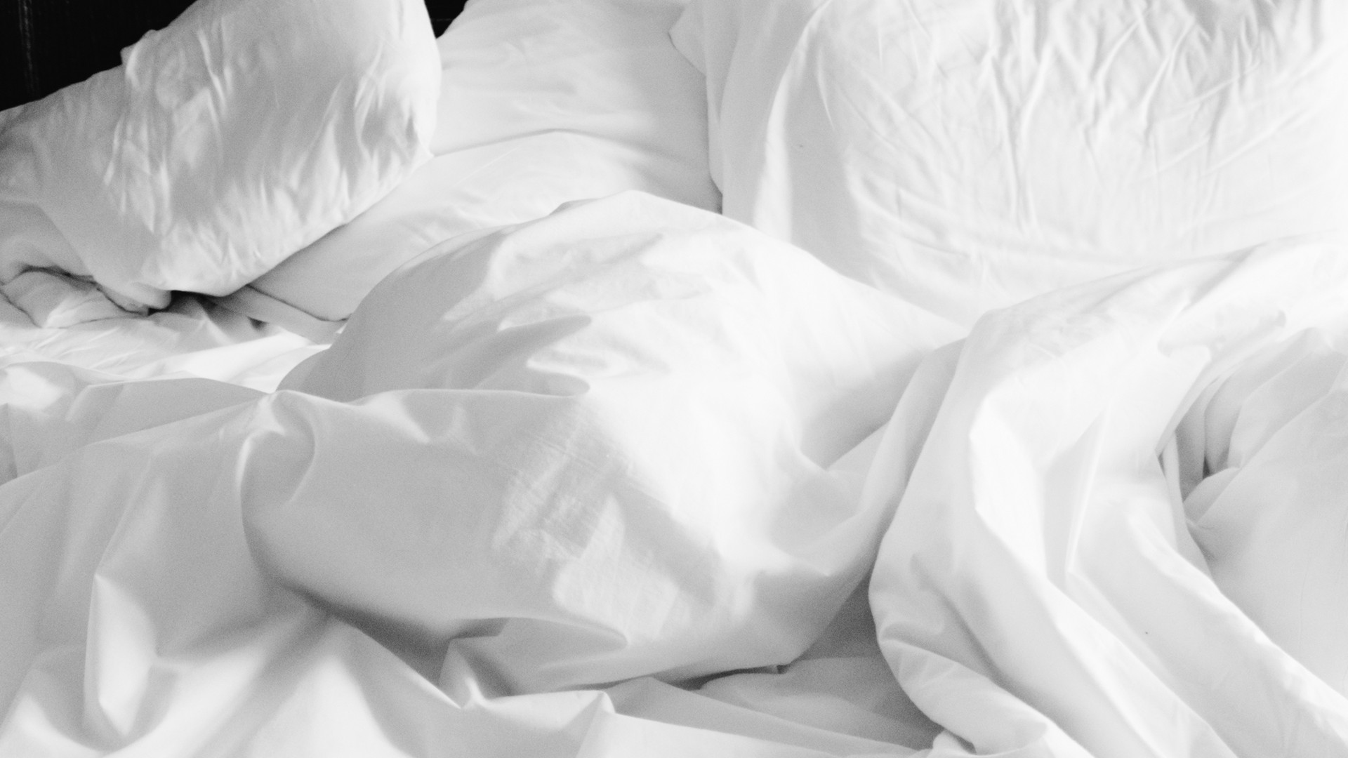 Lits défaits avec draps et oreillers blancs froissés, tête de lit noire, ambiance calme et vide.