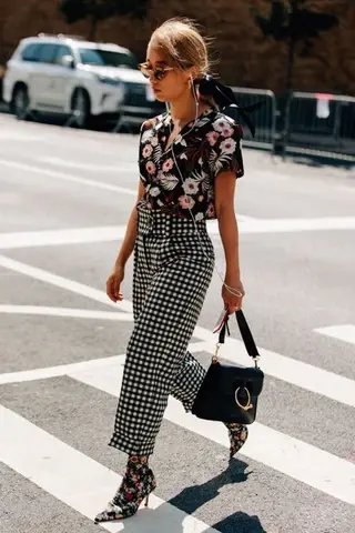 A young woman walks across a crosswalk wearing a floral blouse and checked trousers.