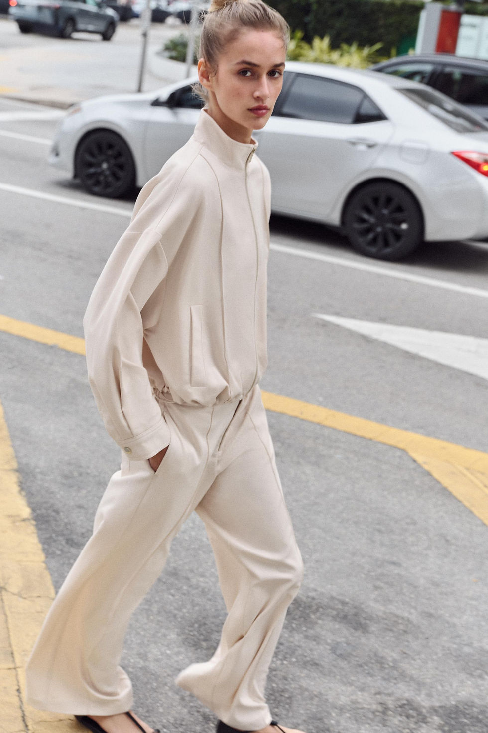 A young girl crossing the street wearing a beige bomber jacket and pants