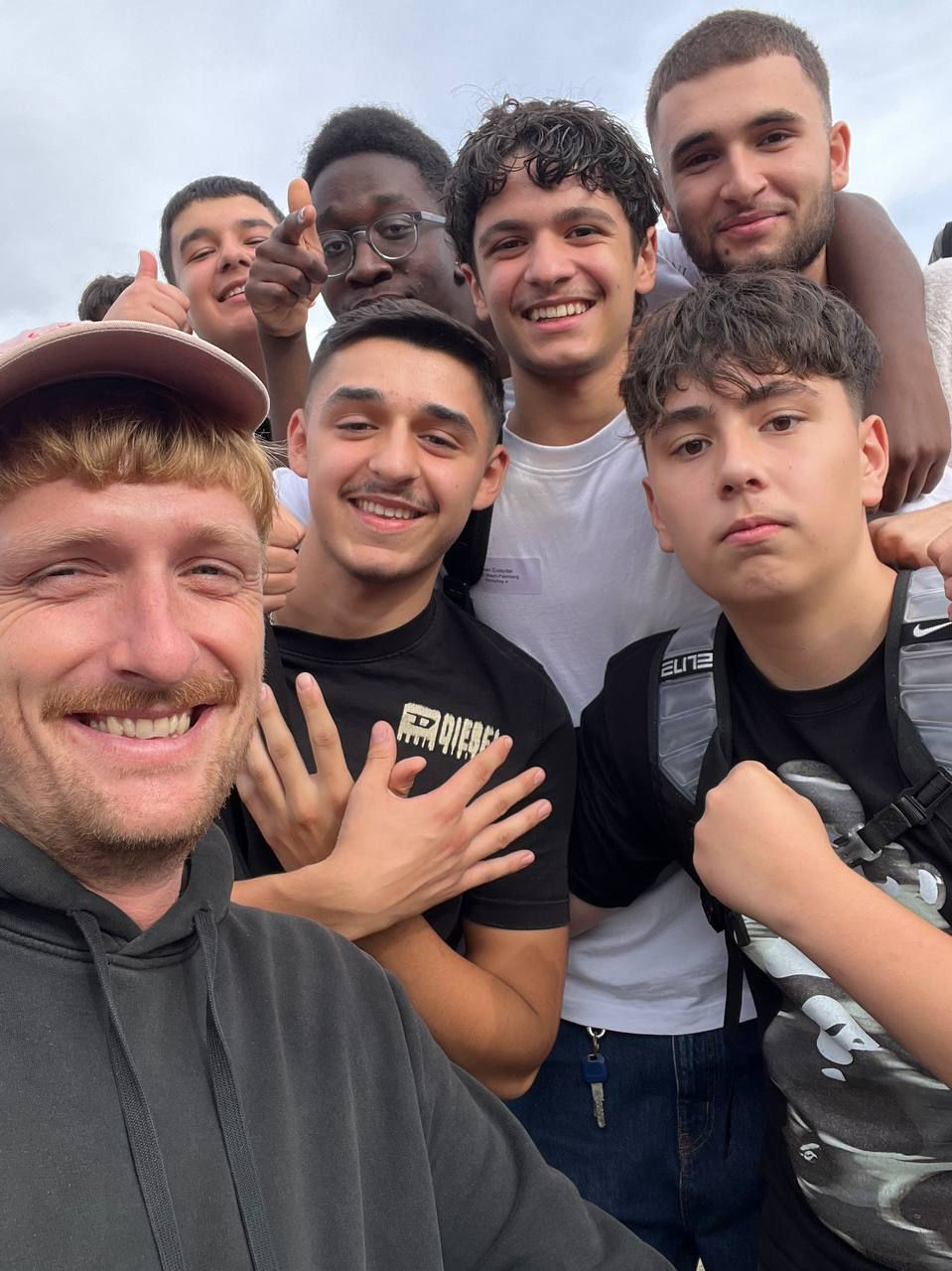 Sechs lächelnde Jungen und ein Mann machen ein Selfie im Freien unter wolkigem Himmel; einer zeigt, andere geben Daumen hoch.