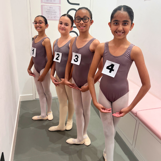 A group of children dressed in matching dance uniforms, each with an examination number pinned to their outfits, stand in the waiting area for their British Ballet Organisation exam at the British Dance Academy in Dubai. They strike their best dance poses, showcasing their preparation and excitement. Their neat uniforms and perfectly styled hair reflect their readiness for the event. The overall mood is a mix of excitement and nervous energy.