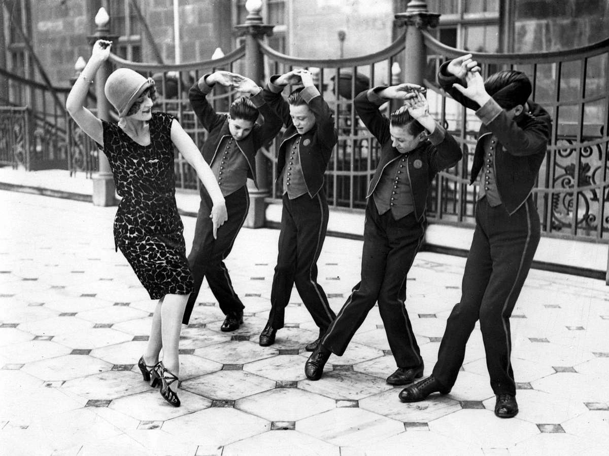 A black-and-white photo from the early 20th century showing a woman and three young boys performing the Charleston dance outdoors. The woman wears a stylish 1920s flapper dress and cloche hat, while the boys are dressed in formal attire with vests and jackets, raising their arms in sync as part of the lively dance