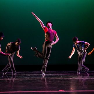 A group of male dancers performing a jazz routine on stage under vibrant lighting. They wear black hats, vests, and pants, with one dancer in the center mid-leap, extending his arm gracefully. The background is dimly lit, emphasizing the dynamic movements and sharp, expressive poses of the dancers.