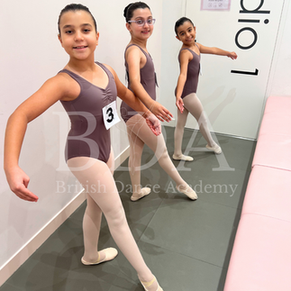 A group of ballet students dressed in matching dance uniforms, each with an examination number pinned to their outfits, standing in the waiting area for their British Ballet Organisation dance exams at British Dance Academy in Dubai. They strike their best dance poses, showcasing their preparation and excitement. Their neat uniforms and perfectly styled hair reflect their readiness for the event. The overall mood is a mix of excitement and nervous energy.