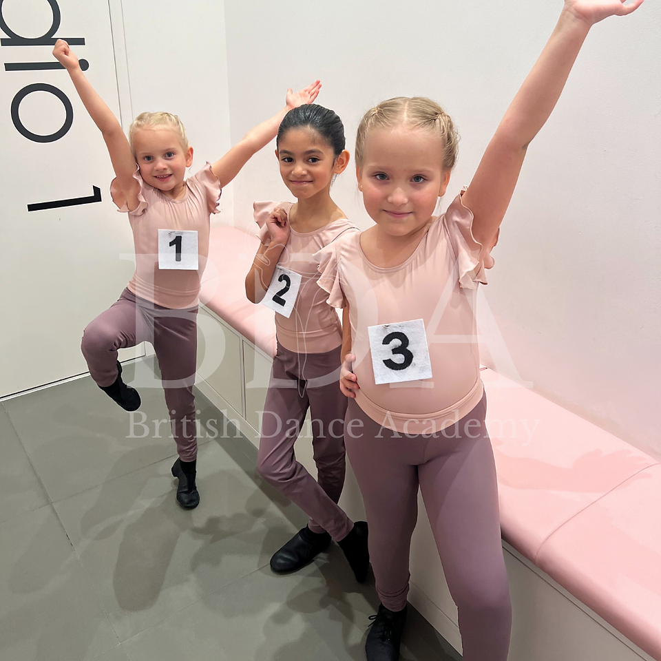 A group of musical theatre dance students dressed in matching dance uniforms, each with an examination number pinned to their outfits, standing in the waiting area for their British Ballet Organisation dance exams at British Dance Academy in Dubai. They strike their best dance poses, showcasing their preparation and excitement. Their neat uniforms and perfectly styled hair reflect their readiness for the event. The overall mood is a mix of excitement and nervous energy.
